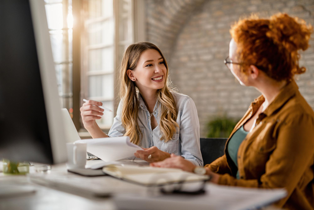 smiling-creative-businesswoman-going-through-paperwork-talking-her-female-colleague-office